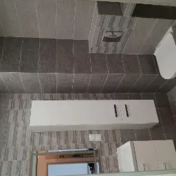 Wykończona łazienka z szarymi płytkami, wiszącą toaletą i białą szafką pod umywalką, widok z dołu ukazujący detale płytek i armatury.