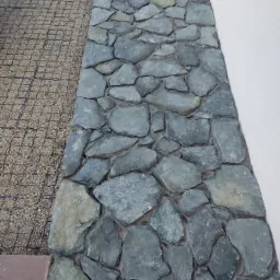 Fragment ściany wykończonej nieregularnymi, szarymi kamieniami, przylegający do białej ściany i powierzchni z drobnym żwirem wzmocnionej siatką.