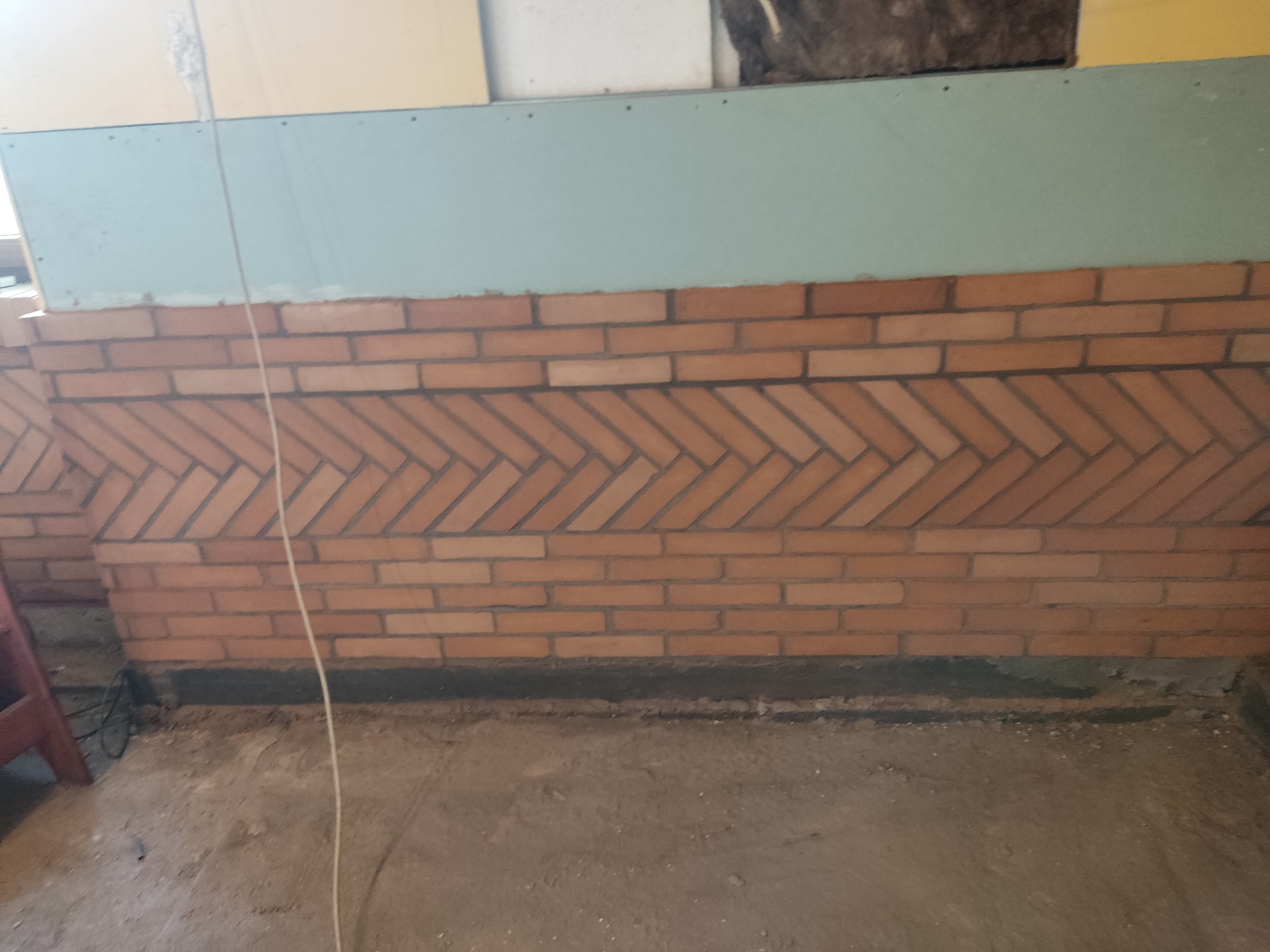 Fragment ściany z cegły klinkierowej ułożonej we wzór jodełki, nad nią płyta gipsowo-kartonowa, widoczne przewody elektryczne i drabina.