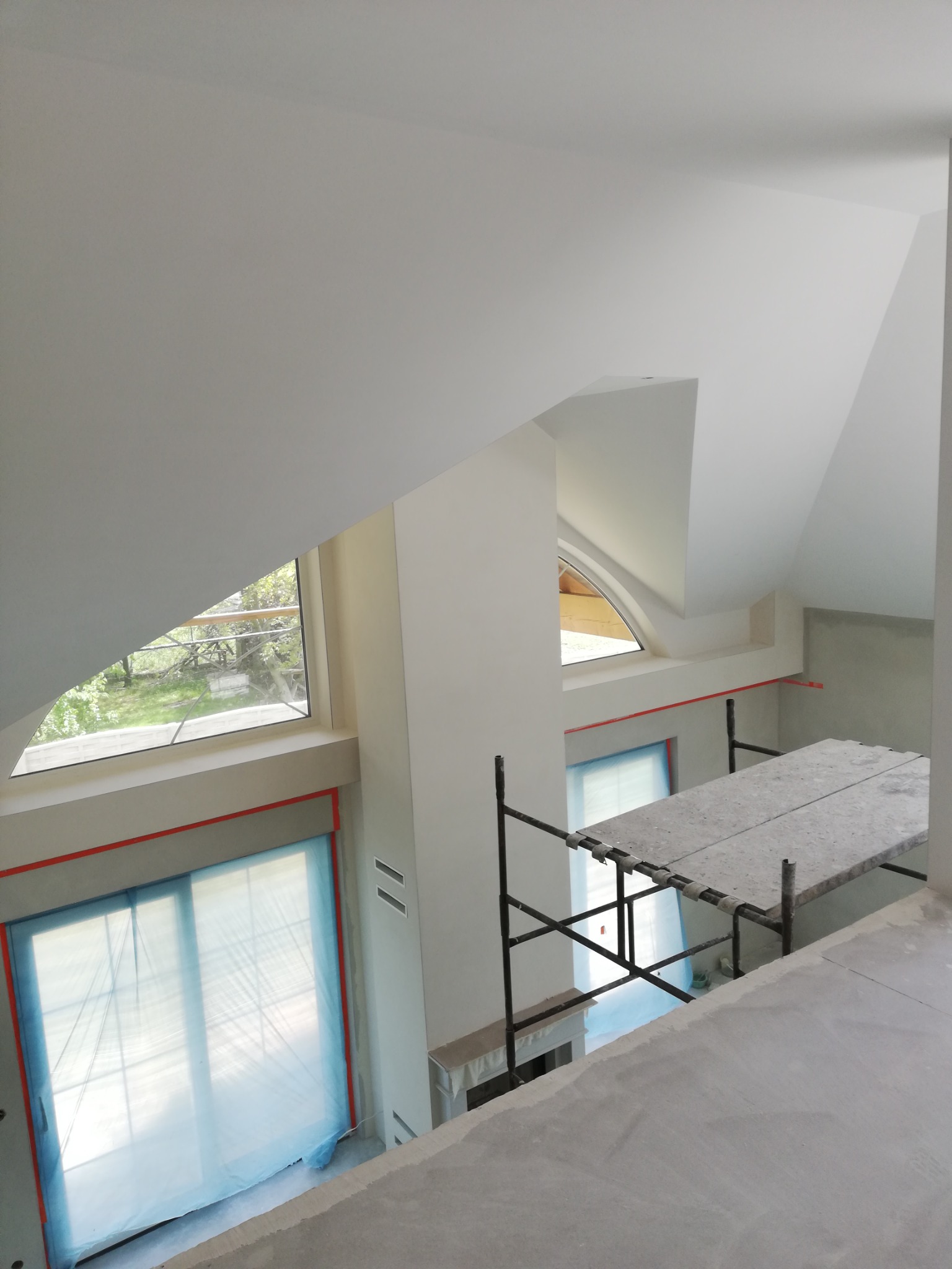 Poddasze w trakcie malowania: widok z góry na rusztowanie, okna zabezpieczone folią, pomalowane na biało ściany o nietypowym kształcie.