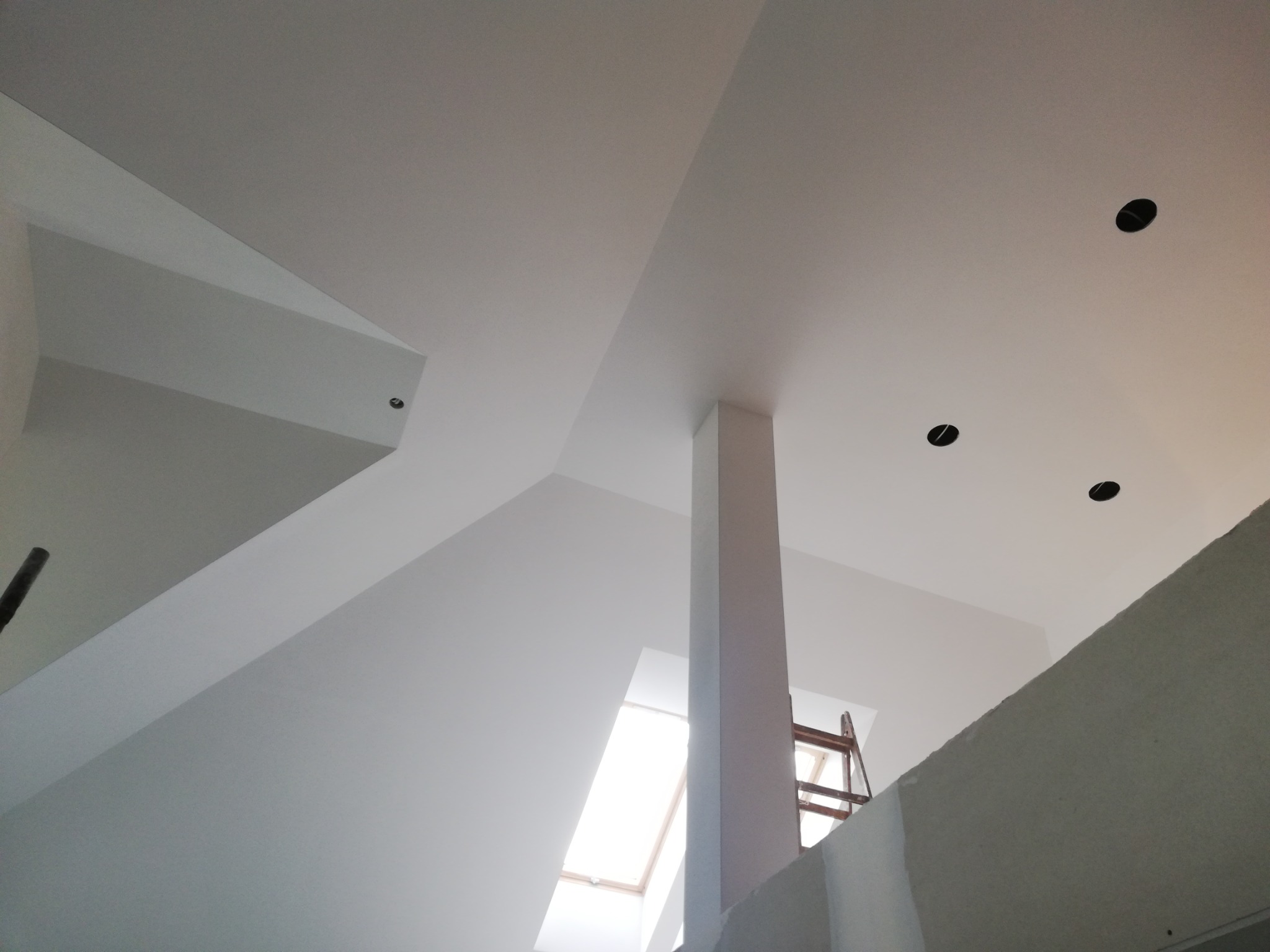 Nowoczesne wnętrze poddasza z białymi ścianami i sufitem, widoczne otwory na oświetlenie punktowe, fragment okna dachowego i drabina malarska w tle.