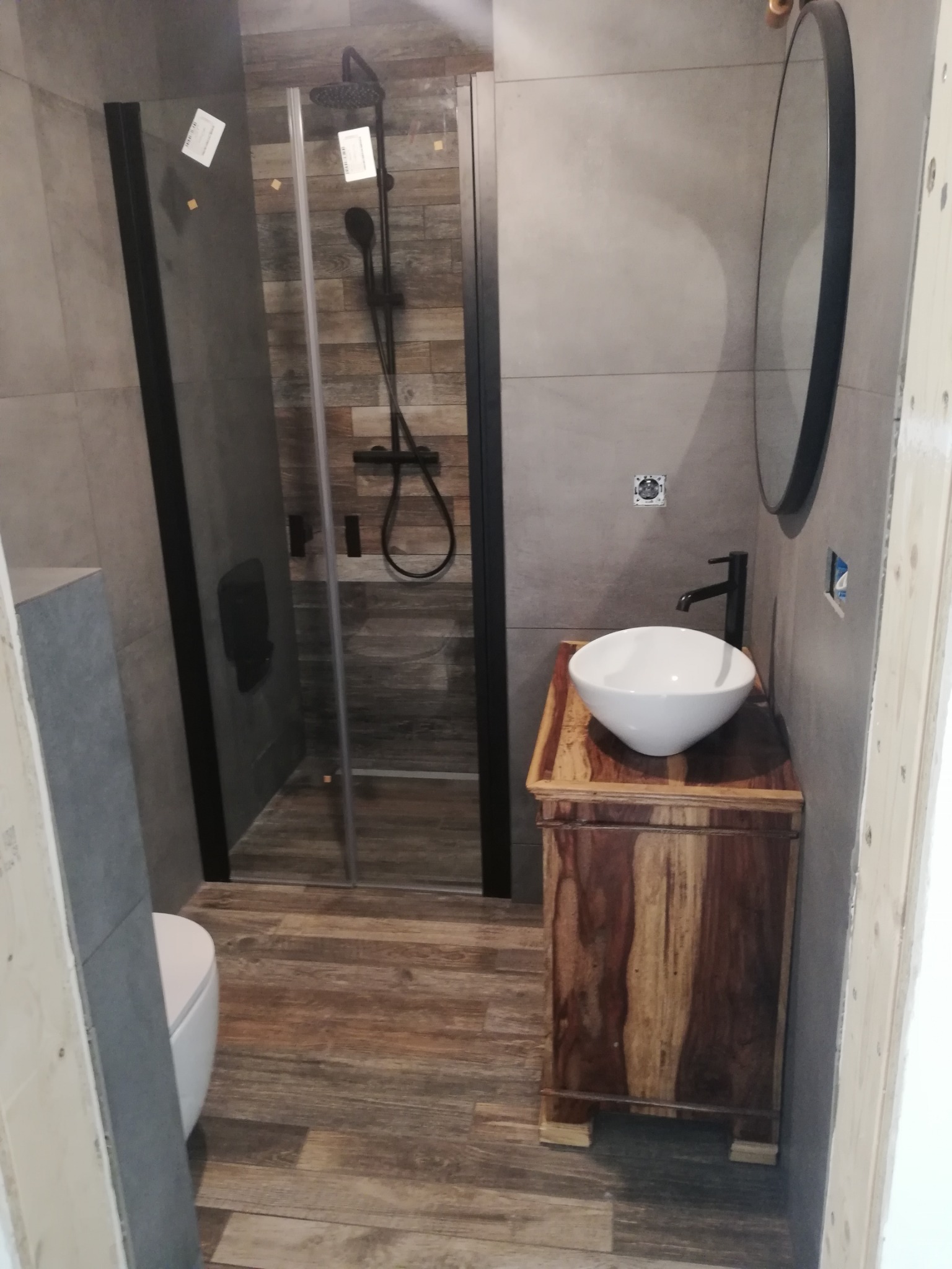 Nowoczesna łazienka z kabiną prysznicową typu walk-in z czarną armaturą, umywalką nablatową na szafce z egzotycznego drewna i okrągłym lustrem w czarnej ramie.