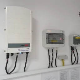 Biała ściana z zamontowanym inwerterem SolarEdge, dwoma skrzynkami elektrycznymi KENO z widocznymi bezpiecznikami, okablowaniem w peszlach oraz fragmentem instalacji rur. Na ścianie widoczne...