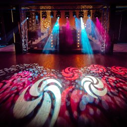 TWENTY ONE- Profesjonalna Obsługa Muzyczna- Jarosław Sitarz - Scena oświetlona reflektorami w kolorach niebieskim i różowym, ze wzorami wyświetlanymi na podłodze, konstrukcja kratowa z reflektorami w tle.