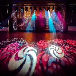 Scena oświetlona reflektorami w kolorach niebieskim i różowym, ze wzorami wyświetlanymi na podłodze, konstrukcja kratowa z reflektorami w tle.