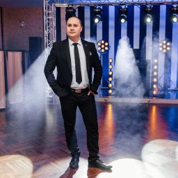 TWENTY ONE- Profesjonalna Obsługa Muzyczna- Jarosław Sitarz - Mężczyzna w czarnym garniturze i białej koszuli z krawatem, stojący na scenie z oświetleniem i lekką mgłą, parkiet z geometrycznym wzorem.