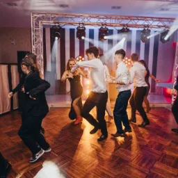 Grupa osób tańczy na parkiecie podczas imprezy, oświetlenie sceniczne z tyłu, parkiet z drewnianych klepek, ujęcie z perspektywy parkietu.
