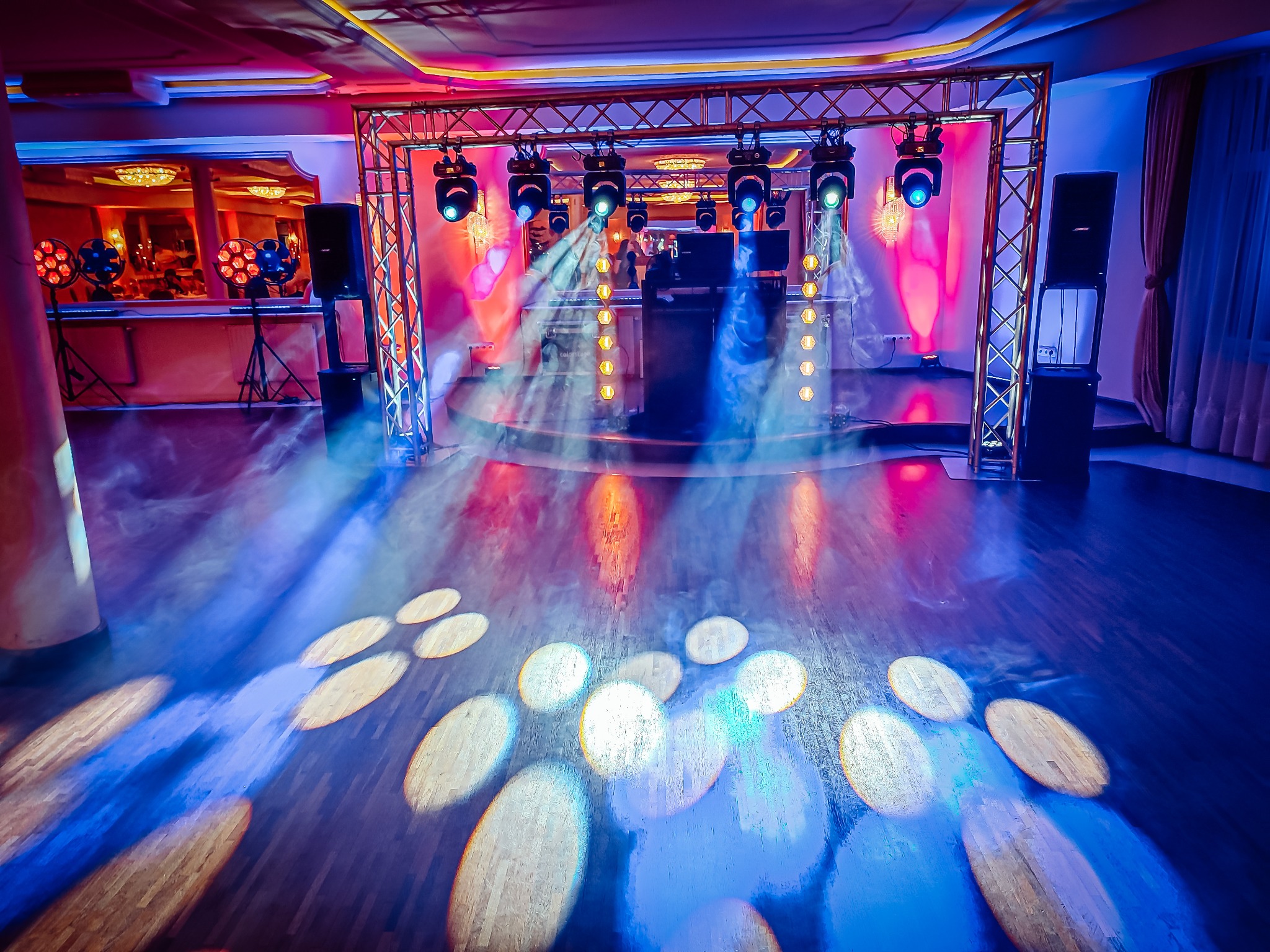 Scena z nagłośnieniem i oświetleniem, reflektory, kolumny głośnikowe, DJ za konsoletą, efekty świetlne na parkiecie, czerwono-niebieska kolorystyka.