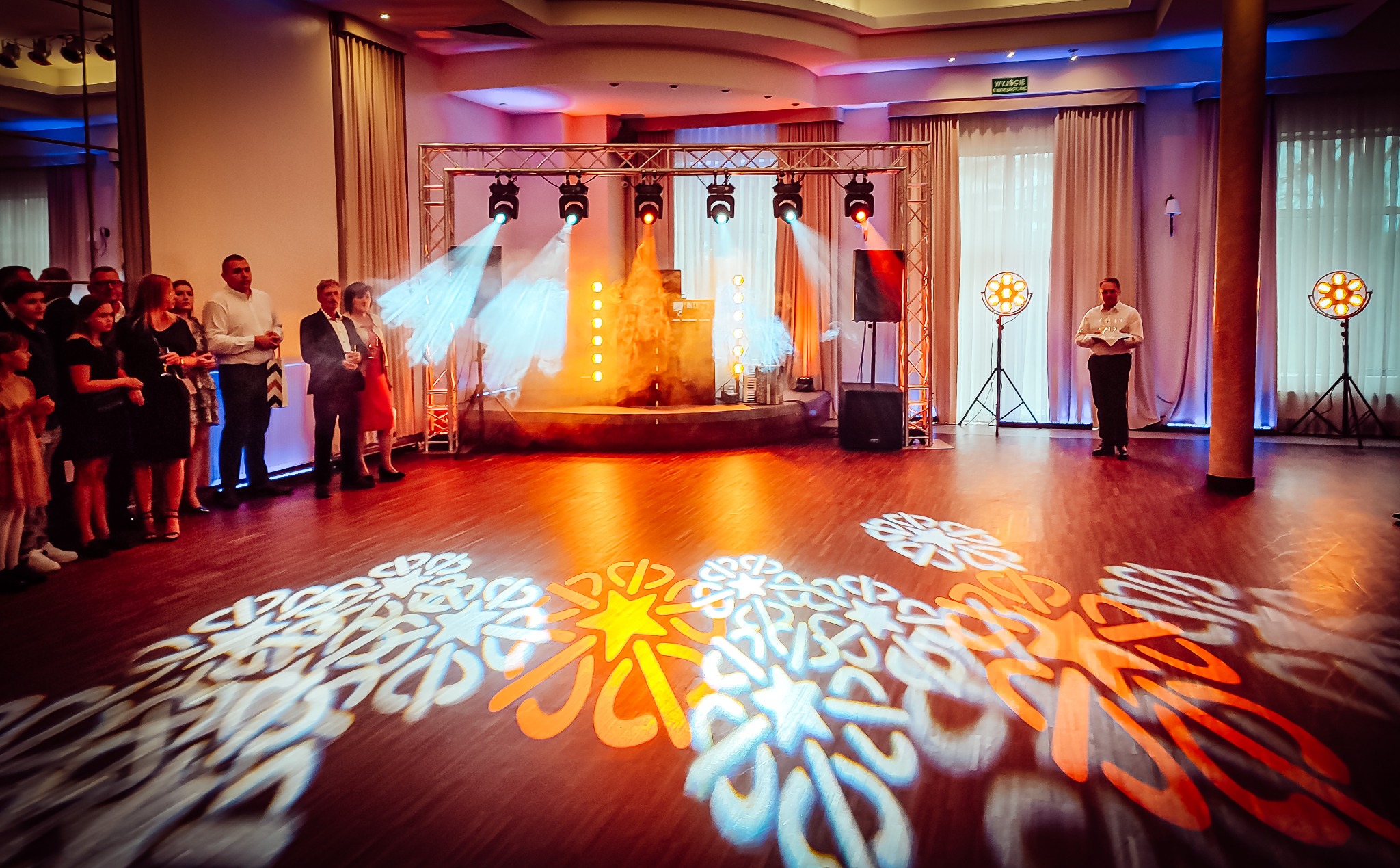 Sala bankietowa z oświetloną sceną, parkietem z projekcjami wzorów i grupą osób obserwujących, mężczyzna z kartką stoi z boku.