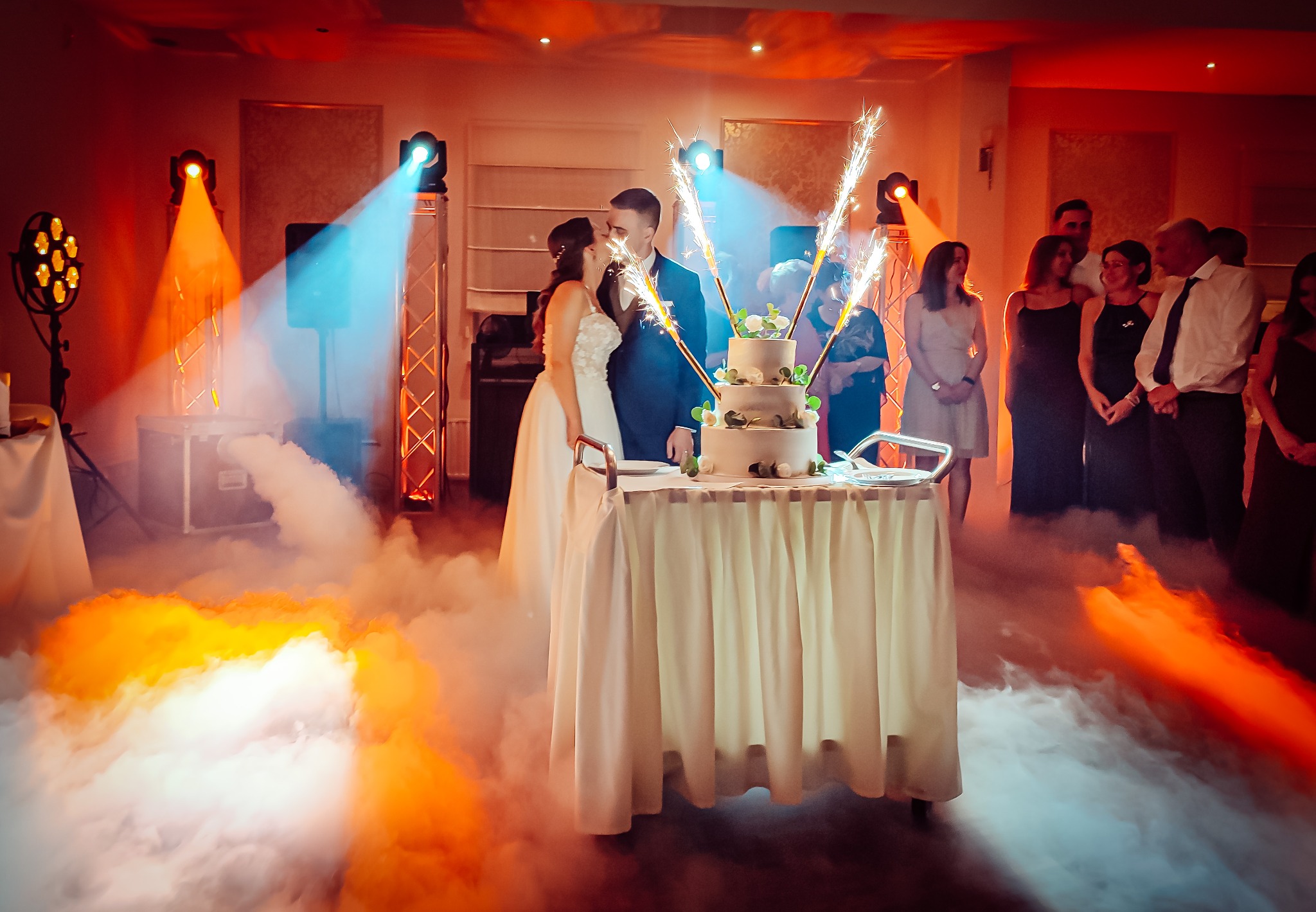 Para młoda całuje się za tortem weselnym z fontannami iskier, oświetlenie sceniczne w tle, dekoracja sali w pomarańczowej mgle.