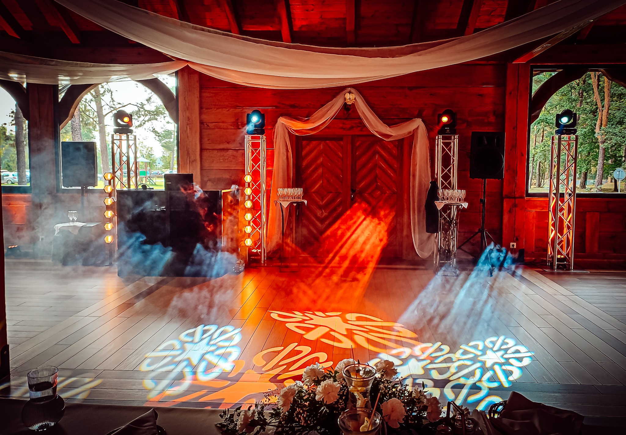 Drewniana sala weselna z dekoracjami świetlnymi, reflektorami na statywach, białymi draperiami i kwiatową dekoracją stołu.
