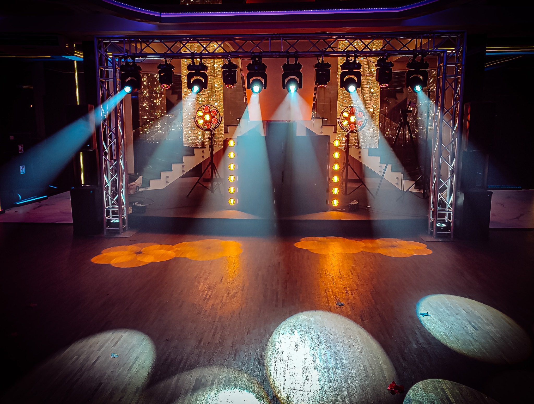 Scena z oświetleniem scenicznym: reflektory punktowe tworzą okrągłe plamy światła na parkiecie, w tle dekoracja z połyskujących elementów i schody.