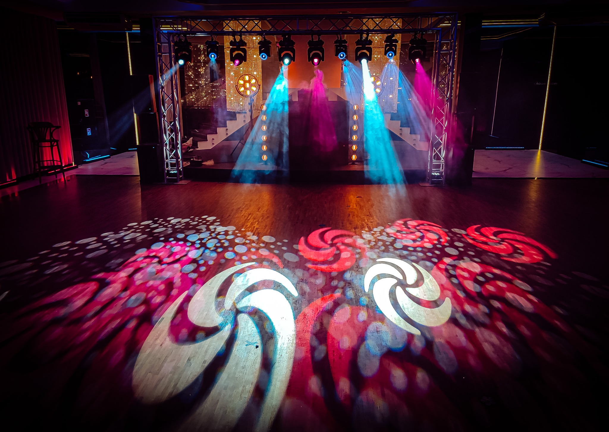 Scena oświetlona reflektorami w kolorach niebieskim i różowym, ze wzorami wyświetlanymi na podłodze, konstrukcja kratowa z reflektorami w tle.