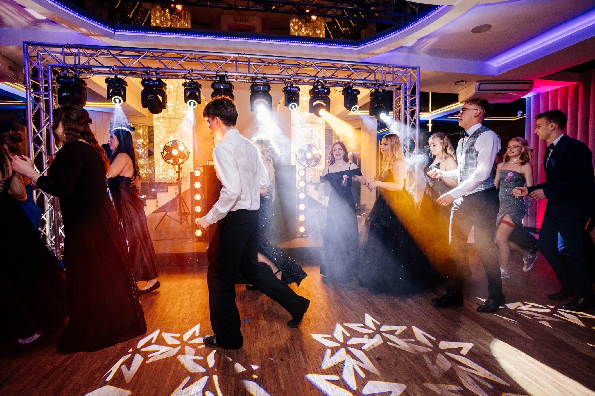 Uczestnicy studniówki w eleganckich strojach tańczą na parkiecie oświetlonym przez reflektory, z widoczną konstrukcją oświetleniową i dekoracyjnym oświetleniem sufitowym.