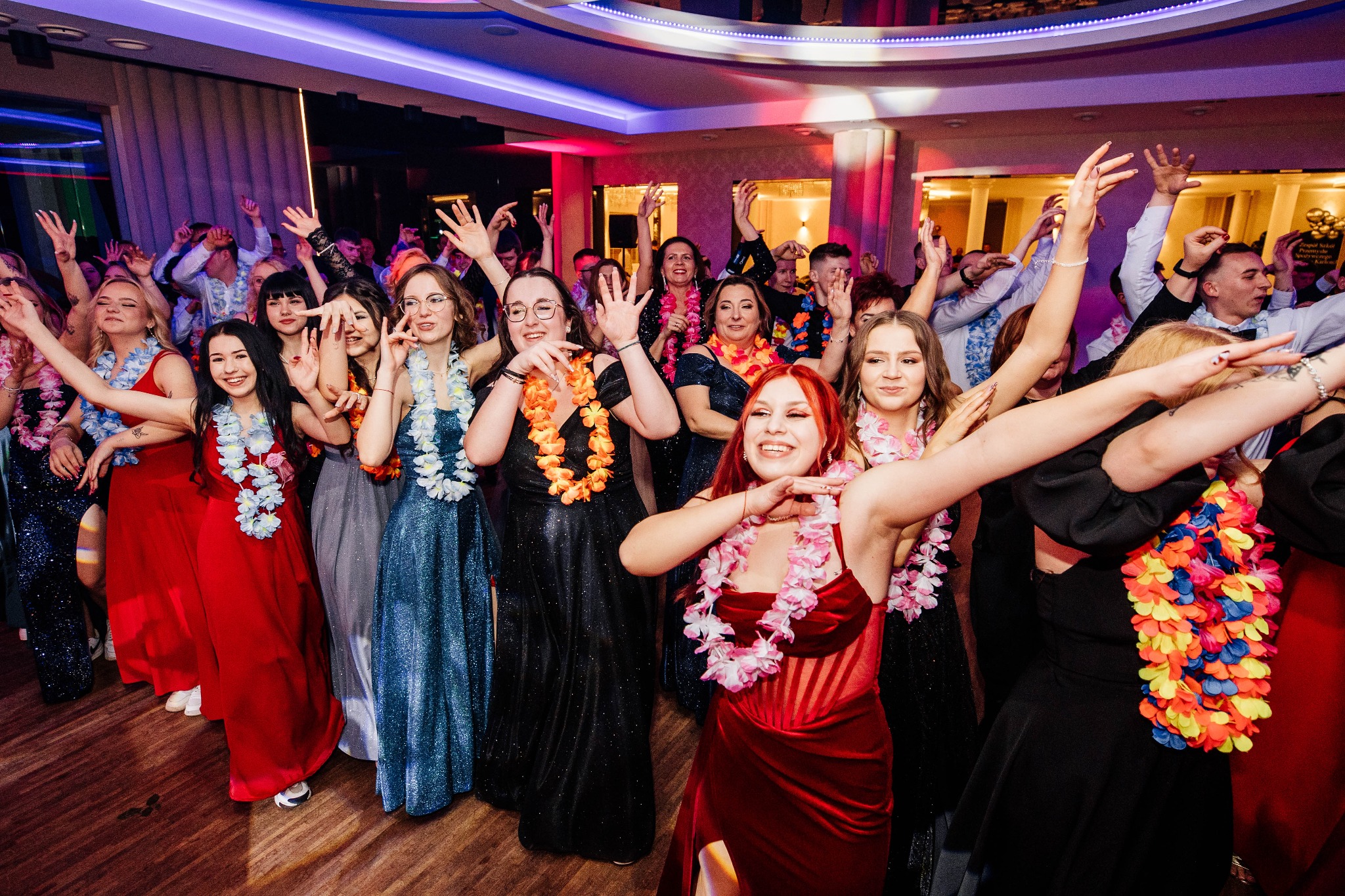 Roztańczona grupa gości w hawajskich naszyjnikach na parkiecie, oświetlenie dyskotekowe, widoczny parkiet i kolumny.