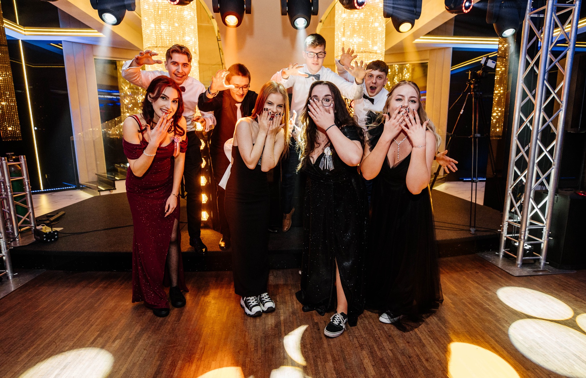 Grupa młodych ludzi w eleganckich strojach pozuje na parkiecie z oświetleniem scenicznym i dekoracyjnym żyrandolem w tle, część osób zakrywa usta dłońmi, wyrażając zaskoczenie lub radość.