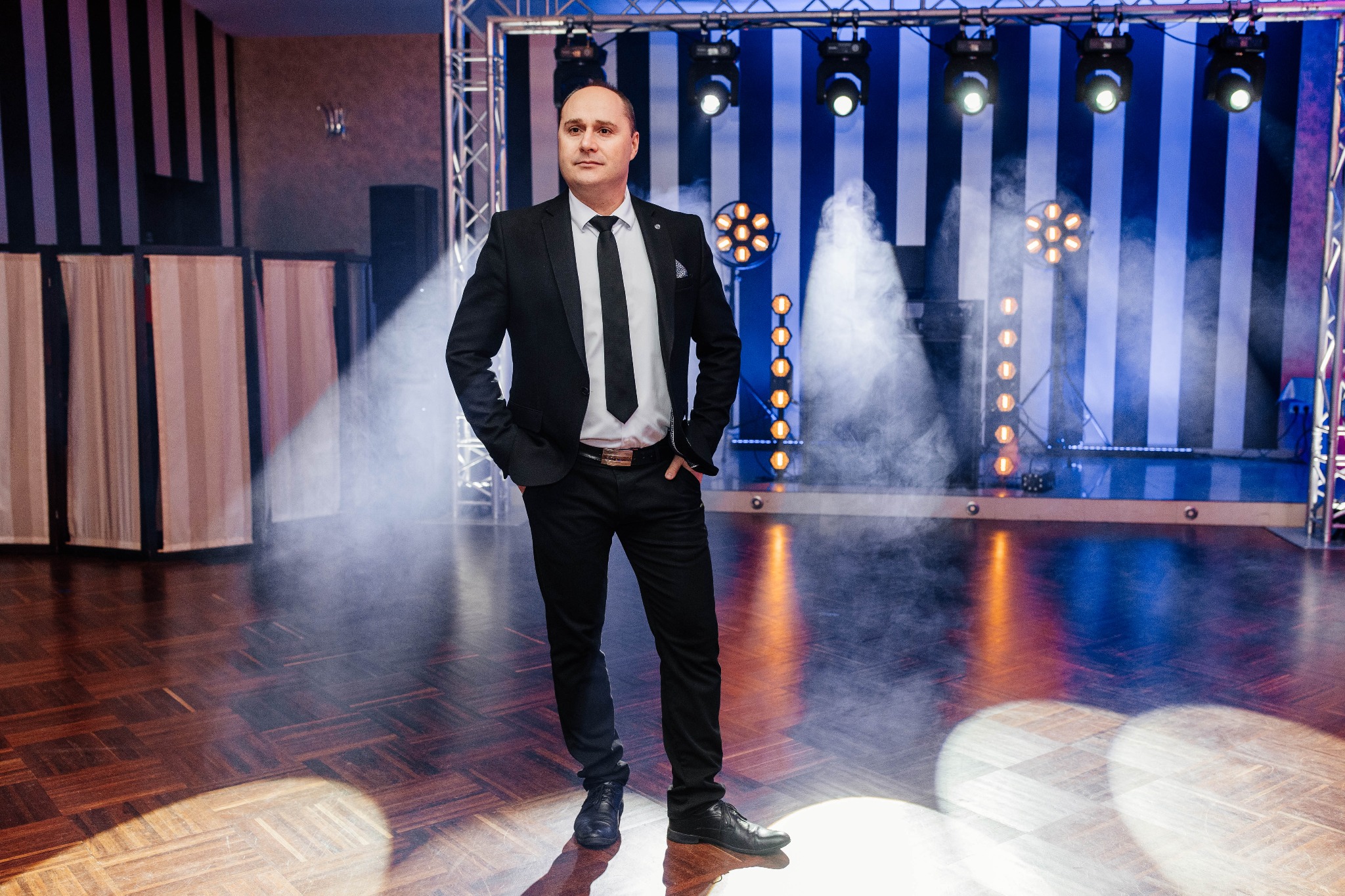 Mężczyzna w czarnym garniturze i białej koszuli z krawatem, stojący na scenie z oświetleniem i lekką mgłą, parkiet z geometrycznym wzorem.