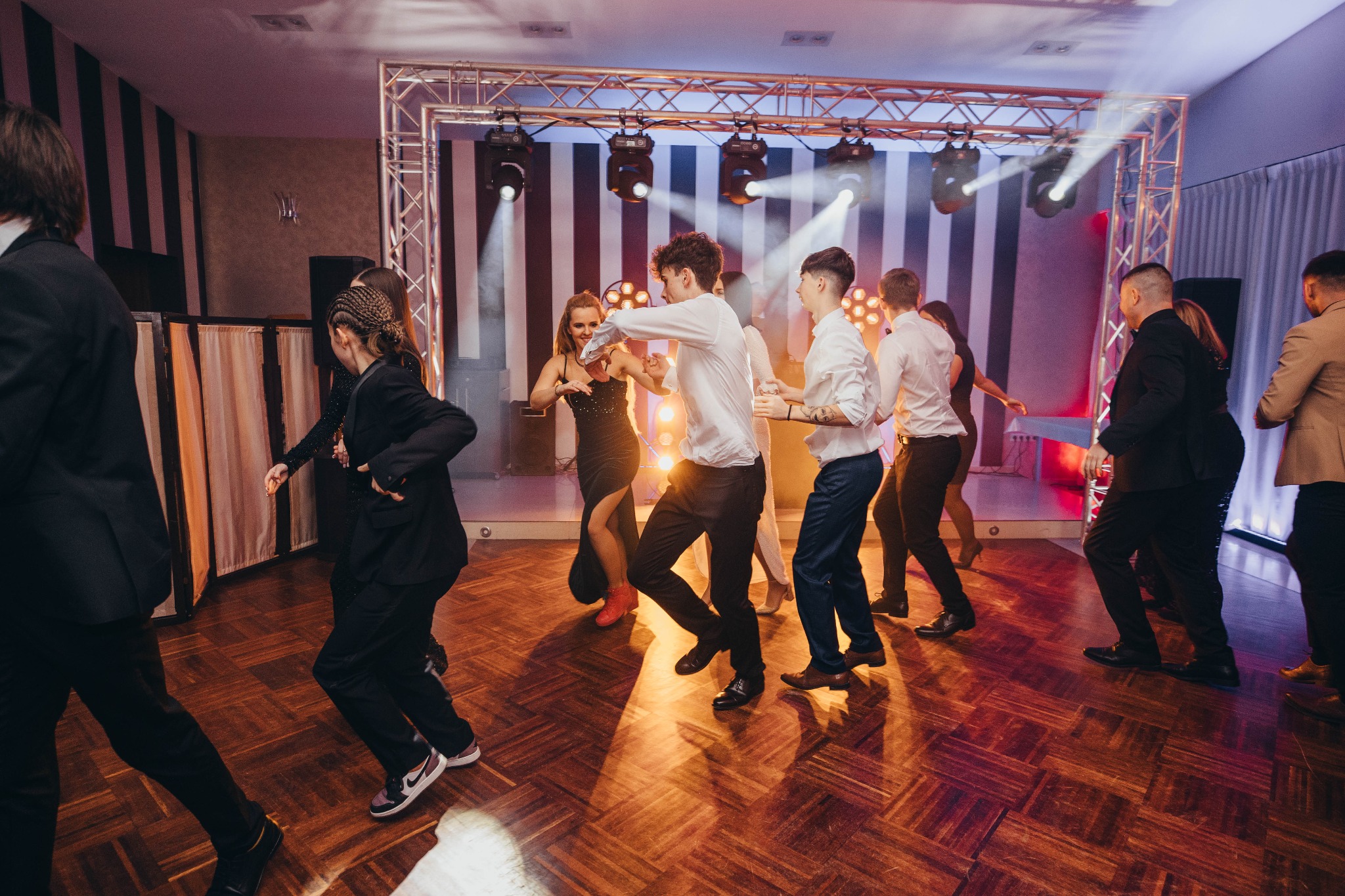 Grupa osób tańczy na parkiecie podczas imprezy, oświetlenie sceniczne z tyłu, parkiet z drewnianych klepek, ujęcie z perspektywy parkietu.