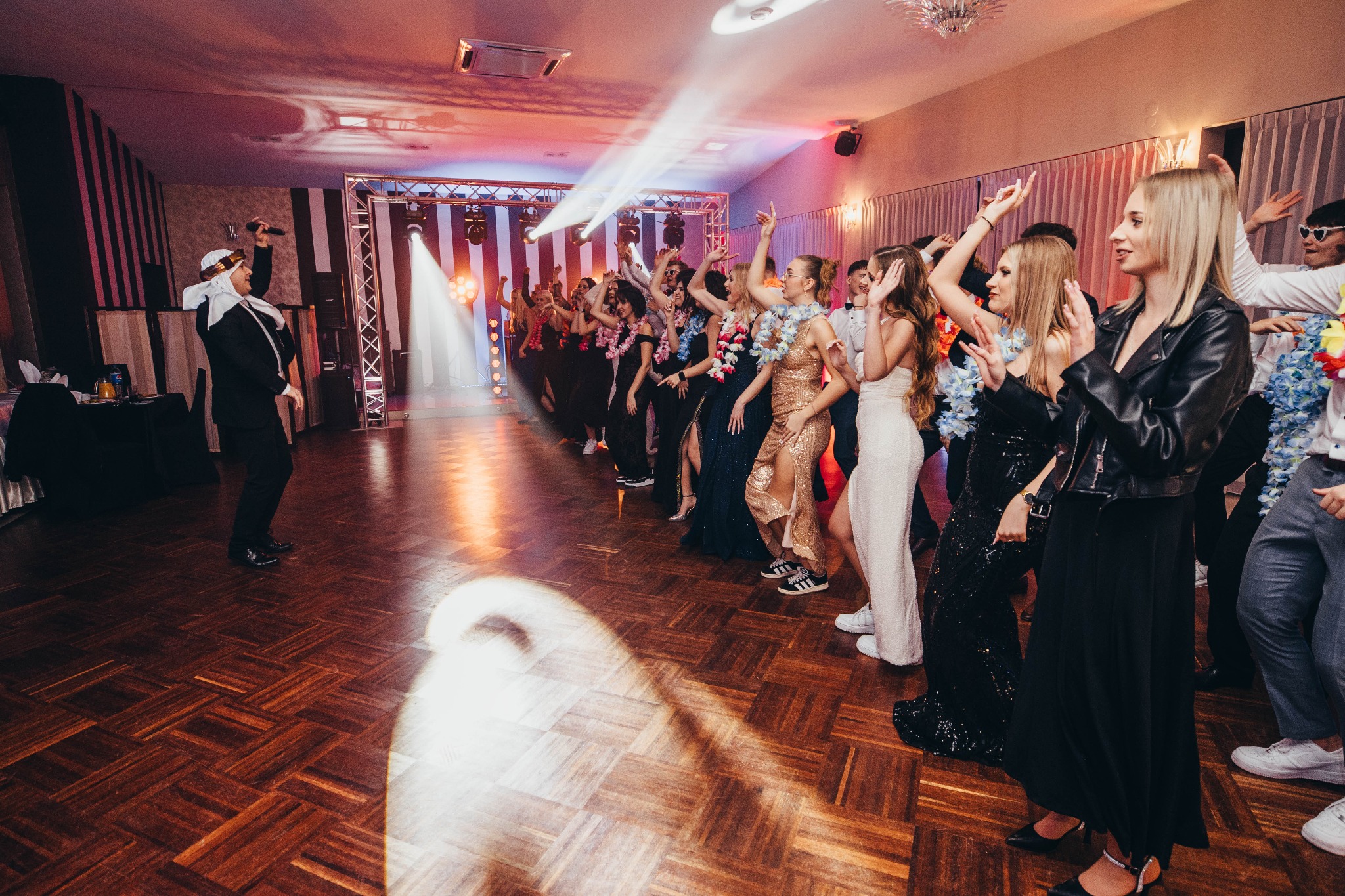 Sala balowa z parkietem i grupą tańczących osób w strojach wieczorowych i kwiatowych girlandach, oświetlona reflektorami. Mężczyzna w garniturze i turbanie na scenie.