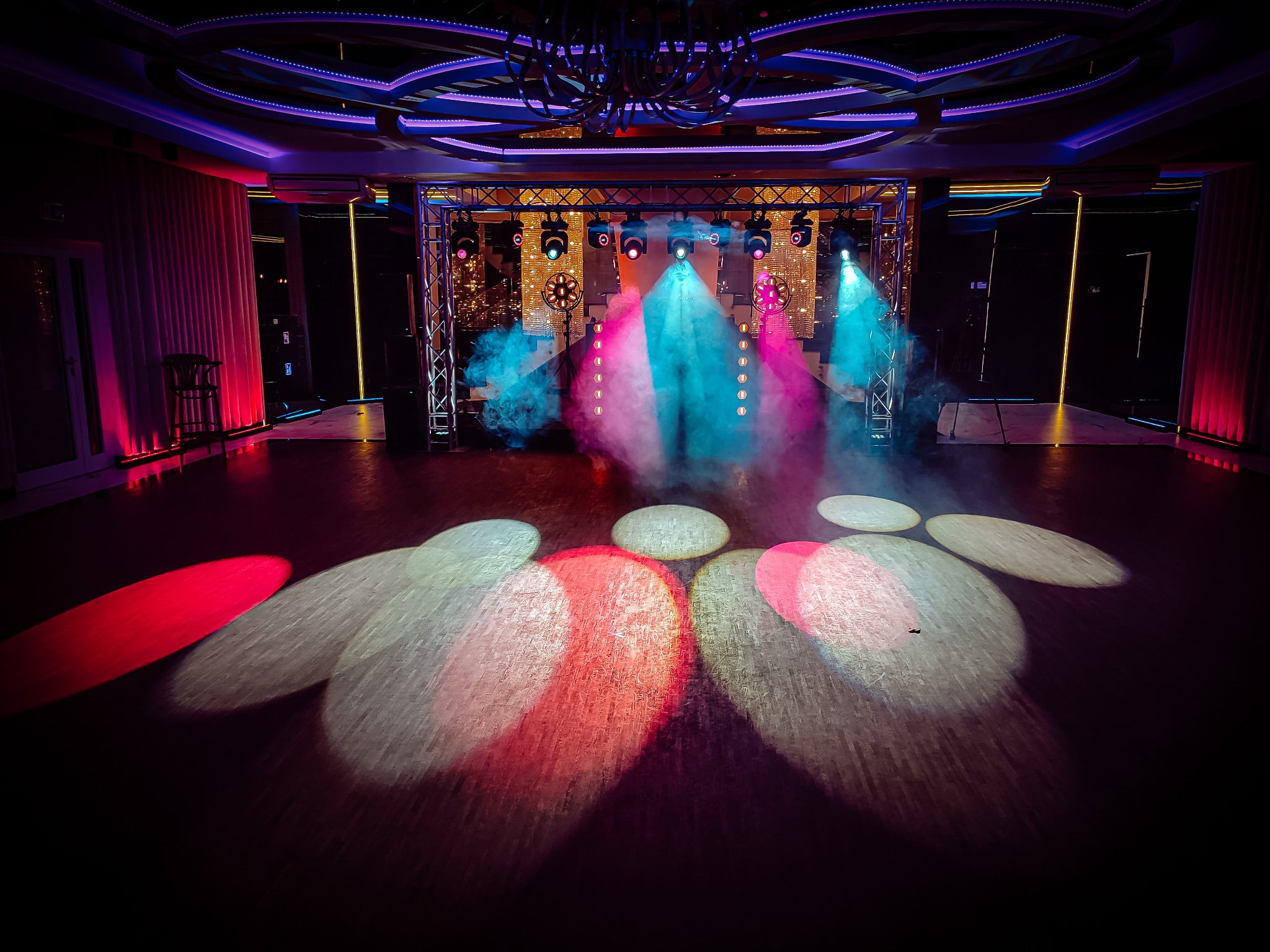 Pusta scena z oświetleniem scenicznym w kolorach różu, błękitu i bieli, z widoczną konstrukcją kratownicową i lekką mgłą w powietrzu.