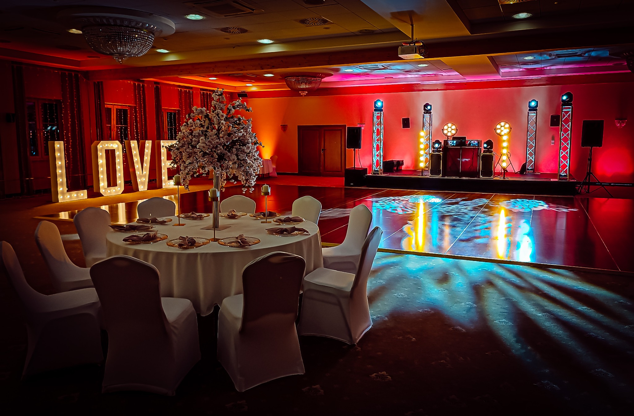 Sala bankietowa z okrągłym stołem, białymi krzesłami, dekoracją kwiatową, podświetlanym napisem LOVE oraz sceną z nagłośnieniem i oświetleniem w czerwono-niebieskich barwach.