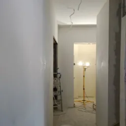 Korytarz w trakcie prac wykończeniowych: ściany pokryte świeżą gładzią, widoczne wiszące kable, drabina malarska, lampa halogenowa na statywie i wiadro z farbą.