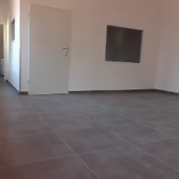 Puste, jasne pomieszczenie z szarymi płytkami podłogowymi, białymi ścianami i drzwiami oraz małym, prostokątnym oknem, ukazujące świeżo położoną podłogę.