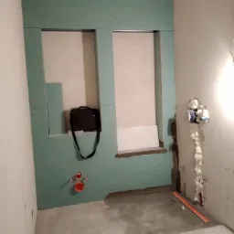 Łazienka w trakcie remontu: zielone płyty kartonowo-gipsowe na ścianie tworzą wnękę, widoczne rury instalacyjne, torba robocza leżąca na półce, ślady pianki montażowej przy rurach.