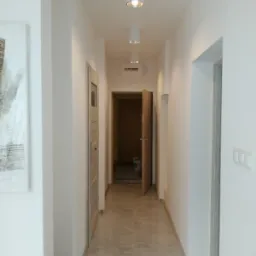 Wykończony korytarz w jasnych barwach z szarą podłogą imitującą marmur, białymi ścianami, trzema okrągłymi lampami sufitowymi i dwiema parami drzwi po bokach, z widoczną skrzynką elektryczną...