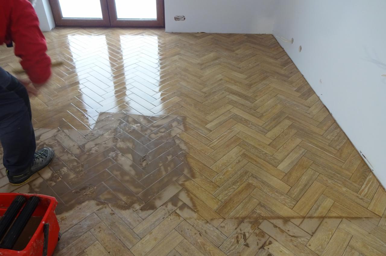 Lakierowanie parkietu ułożonego w jodełkę, widoczna różnica między polakierowaną a surową częścią podłogi, czerwone wiaderko z wałkami w rogu.