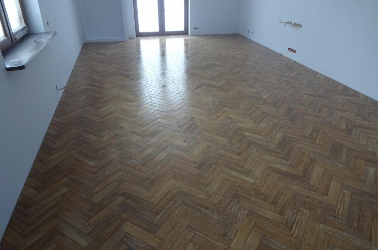 Ułożona parkietem w jodełkę podłoga w jasnym, pustym pokoju z białymi ścianami i oknem balkonowym w tle.