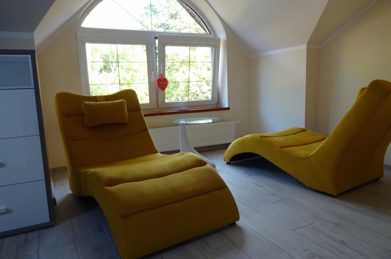 Jasne wnętrze poddasza z dwoma żółtymi leżankami relaksacyjnymi, małym okrągłym stolikiem i oknem z widokiem na zieleń.