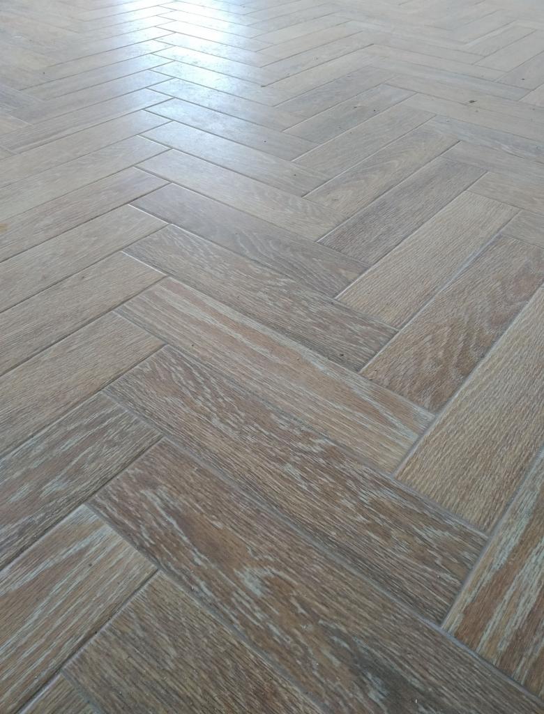 Jasnobrązowa podłoga z płytek ułożonych w jodełkę, imitujących drewno z widoczną strukturą słojów, lekko odbijająca światło.