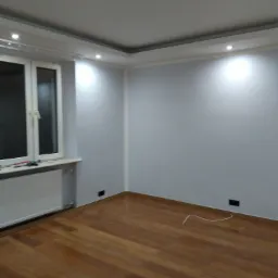 Pomalowane na szaro wnętrze pokoju z drewnianą podłogą, białym oknem i grzejnikiem oraz dekoracyjnym oświetleniem sufitowym.