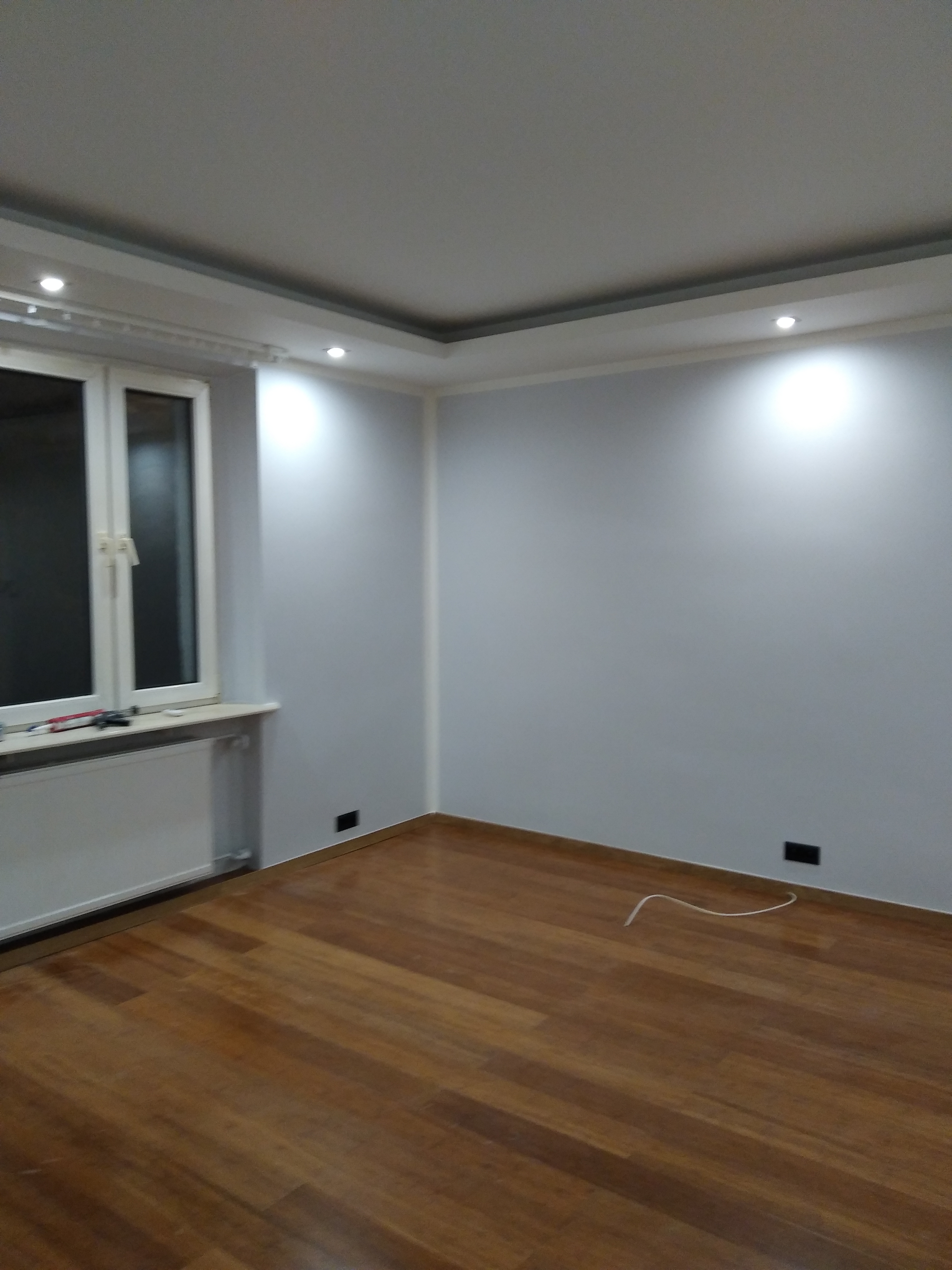 Pomalowane na szaro wnętrze pokoju z drewnianą podłogą, białym oknem i grzejnikiem oraz dekoracyjnym oświetleniem sufitowym.