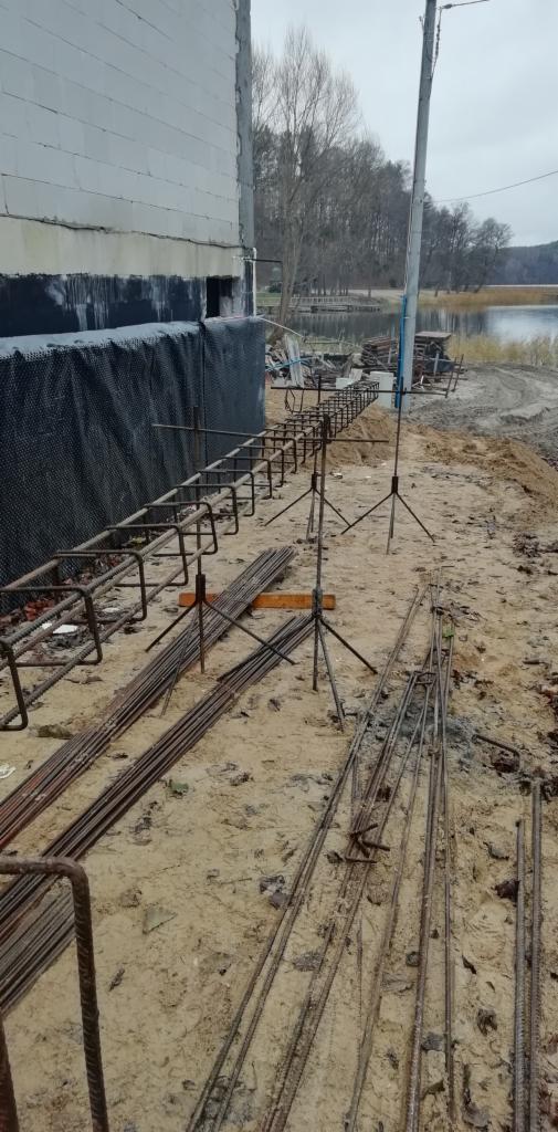 Przygotowanie zbrojenia fundamentów na piaszczystym gruncie, widoczny budynek w trakcie izolacji, w tle jezioro i pochmurne niebo.