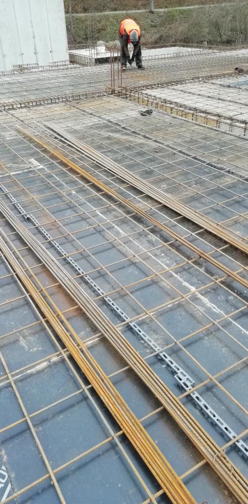 Prace zbrojarskie na budowie: siatka zbrojeniowa ułożona na czarnej folii, w tle pracownik w pomarańczowej kamizelce.