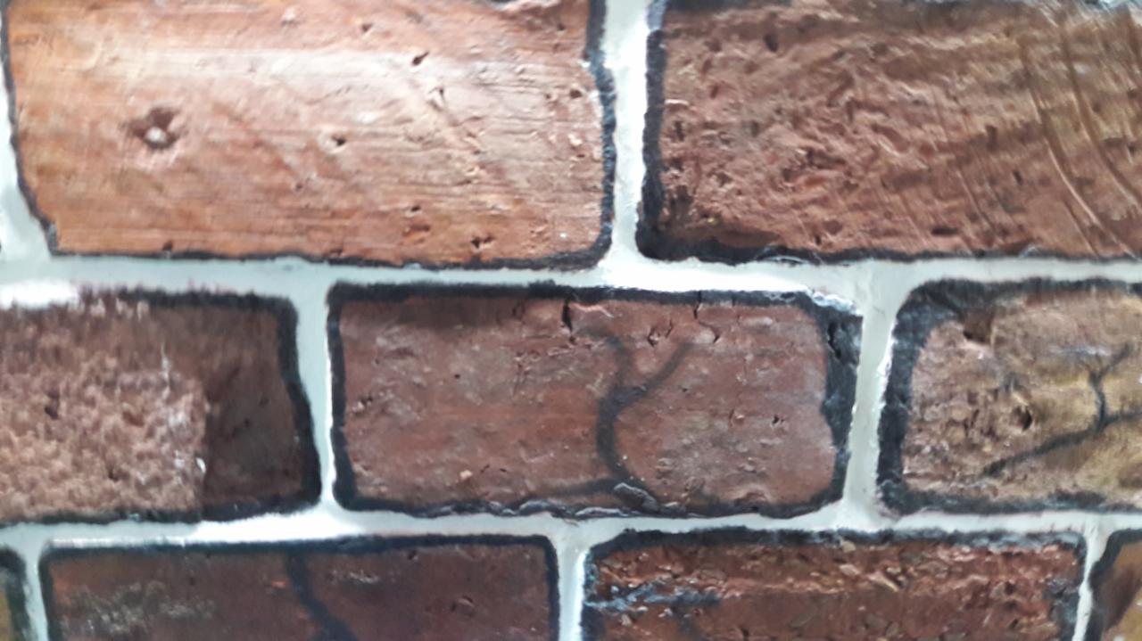 Fragment ściany wyłożonej cegłami w odcieniach czerwieni i brązu, z wyraźnie zaznaczonymi fugami w kolorze jasnoszarym i czarnym obrysem, imitującymi starą cegłę.