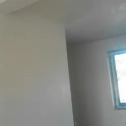 Świeżo położony, gładki tynk na ścianach wewnętrznych pomieszczenia, widoczna listwa sufitowa i wiadro z farbą w rogu.