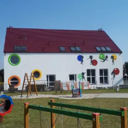Biały budynek przedszkola z czerwonym dachem, okrągłymi oknami w kolorowych obwódkach i rysunkami dzieci na elewacji, plac zabaw w pierwszym planie.