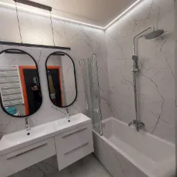 Wykończona łazienka z białymi marmurowymi płytkami, owalnymi lustrami z czarną ramą, białą szafką pod umywalkę i wanną z parawanem, oświetlona listwami LED.