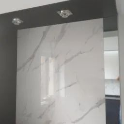 Ściana wyłożona wielkoformatowymi płytkami imitującymi marmur Carrara, z minimalistycznym oświetleniem punktowym w suficie podwieszanym, obok pionowy pas lustra.