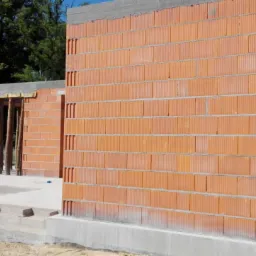 Wznoszenie ścian z pustaków ceramicznych na fundamencie z betonu, widoczne drewniane stemple podtrzymujące strop, zielone drzewa w tle.