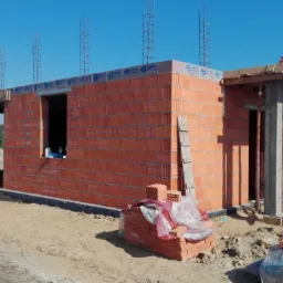 Wznoszenie ścian domu z czerwonej cegły ceramicznej Porotherm, widoczne zbrojenia pionowe i podpora z drewnianych bali, na pierwszym planie sterta cegieł i narzędzia.