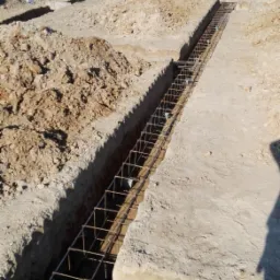 Zbrojenie ławy fundamentowej ułożone w wykopie, widok z góry na plac budowy z hałdami ziemi i zielenią w tle.