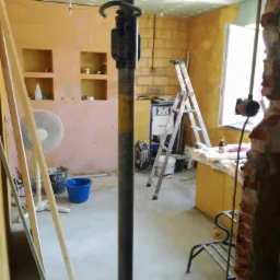 Wnętrze w trakcie remontu z widocznym stemplem budowlanym, ścianami przygotowanymi pod szpachlowanie, drabina aluminiowa, wentylator i narzędzia malarskie.