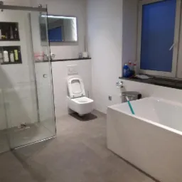 Nowoczesna łazienka w odcieniach bieli i szarości z kabiną prysznicową typu walk-in, podwieszaną toaletą i wanną pod oknem.