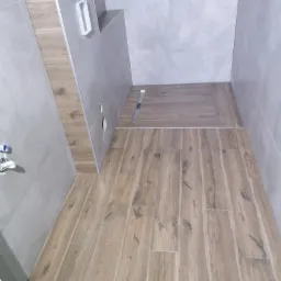 Wykończona łazienka z drewnopodobnymi płytkami podłogowymi i szarymi płytkami ściennymi, widoczny odpływ liniowy i zabudowa pod umywalkę z elementami drewnianymi.