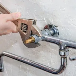 Regulowany klucz nasadowy dokręca mosiężną złączkę na chromowanym kranie zamontowanym na białej, teksturowanej ścianie.