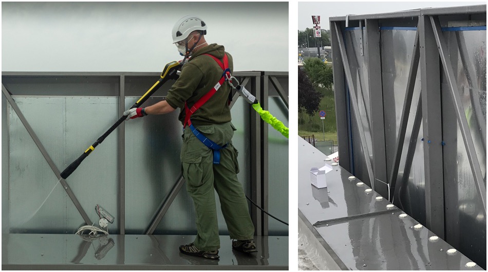 Pracownik w uprzęży, kasku i masce ochronnej myje elewację budynku za pomocą myjki ciśnieniowej Karcher, widoczne odbicia na mokrej powierzchni.